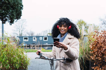 Cheerful young African American female with curly dark hair in warm coat riding bicycle while using mobile phone and listening to music in headphones in autumn day - ADSF33775