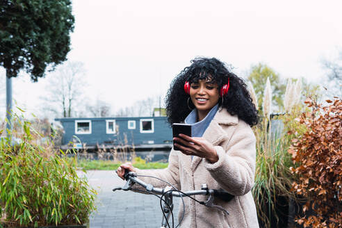 Fröhliche junge Afroamerikanerin mit lockigem dunklem Haar in warmem Mantel fährt Fahrrad, während sie ein Mobiltelefon benutzt und über Kopfhörer Musik hört, an einem Herbsttag - ADSF33775