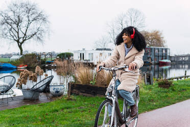 Full body of cheerful young African American female with curly hair in warm clothes riding bicycle while listening to music in headphones in autumn day and looking away - ADSF33776