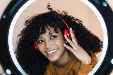 Cheerful young African American female with curly dark hair listening to music in headphones illuminated by ring lamp and looking away against white wall - ADSF33779