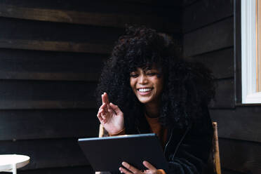 Fröhliche junge Afroamerikanerin in warmer Jacke surft auf einem Tablet, während sie auf einem Stuhl auf der Terrasse eines Hauses sitzt und auf den Bildschirm schaut - ADSF33780