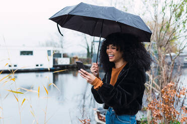 Seitenansicht einer entzückten jungen Afroamerikanerin in trendiger, warmer Kleidung, die auf ihrem Handy surft, während sie mit einem Regenschirm an einem regnerischen Tag steht - ADSF33781