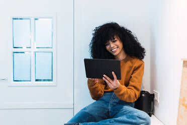 Lächelnde junge Afroamerikanerin mit lockigen dunklen Haaren, die Jeans und Sweatshirt trägt, surft auf einem Tablet, während sie auf dem Küchentisch sitzt und auf den Bildschirm vor der weißen Wand schaut - ADSF33782