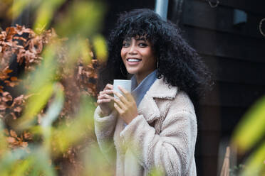 Positive junge Afroamerikanerin mit lockigem dunklem Haar im warmen Mantel, die ein heißes Getränk aus einem Becher trinkt, während sie draußen in der Nähe eines Hauses steht und wegschaut - ADSF33783