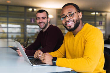 Lächelnde, multirassische, bärtige Partner schauen in die Kamera, während sie zusammen an einem Tisch mit Laptop und Tablet in einem Coworking Center sitzen - ADSF33784