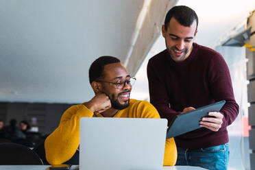 Lächelnde, multirassische, bärtige Partner arbeiten gemeinsam an einem Projekt an einem Tisch mit Laptop und Tablet in einem Coworking Center - ADSF33786