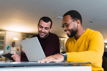 Lächelnde, multirassische, bärtige Partner arbeiten gemeinsam an einem Projekt an einem Tisch mit Laptop und Tablet in einem Coworking Center - ADSF33788