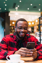 Smiling African American bearded male in red checkered shirt messaging on mobile phone wit earphones while drinking coffee in cafe and looking at screen - ADSF33790