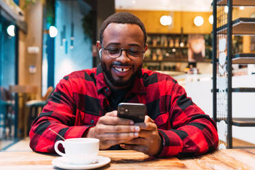 Smiling African American bearded male in red checkered shirt messaging on mobile phone wit earphones while drinking coffee in cafe and looking at screen - ADSF33791