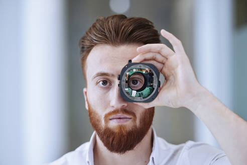 Junger Mann schaut durch ein offenes Kameraobjektiv in einem Büro - CVF01955
