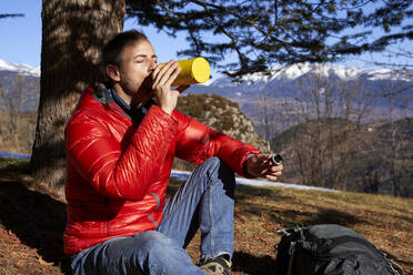 Man drinking water from bottle sitting under tree on sunny day - VEGF05293