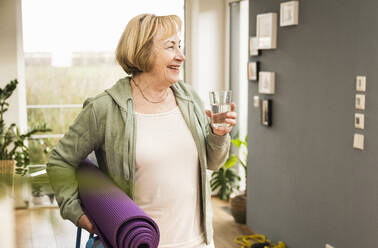 Glückliche ältere Frau mit Übungsmatte, die ein Glas Wasser zu Hause hält - UUF25655