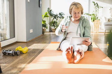 Smiling senior using digital tablet PC sitting on exercise mat at home - UUF25664