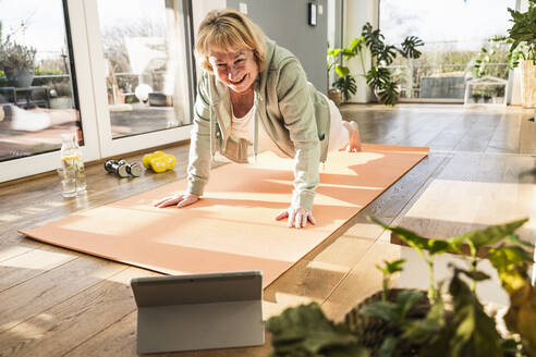 Glückliche ältere Frau, die mit Hilfe eines Online-Tutorials auf einem Tablet-PC zu Hause die Planke übt - UUF25667