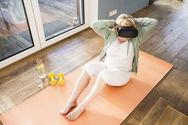 Ältere Frau mit Virtual-Reality-Simulator, die zu Hause mit den Händen hinter dem Kopf trainiert - UUF25680
