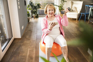 Blonde ältere Frau, die ein Smartphone benutzt und mit einer Hantel auf einem Fitnessball zu Hause trainiert - UUF25682