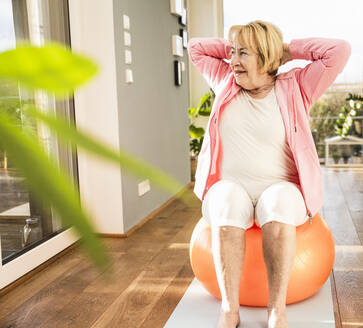 Ältere Frau sitzt mit den Händen hinter dem Kopf auf einem Fitnessball zu Hause - UUF25684