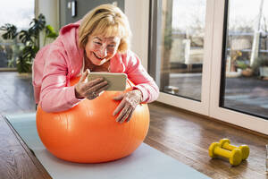 Glückliche ältere Frau mit Smartphone beim Training auf einem Fitnessball zu Hause - UUF25689