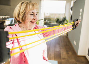 Happy senior woman doing stretching exercise with resistance band at home - UUF25693