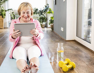 Glückliche ältere Frau mit Tablet-PC, die zu Hause auf einer Übungsmatte sitzt - UUF25696