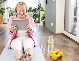 Happy senior woman with tablet PC sitting on exercise mat at home - UUF25696