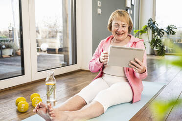 Happy blond senior woman looking at tablet PC on exercise mat at home - UUF25698