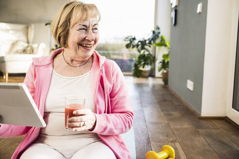 Cheerful senior woman with glass of juice sitting on ground at home - UUF25700
