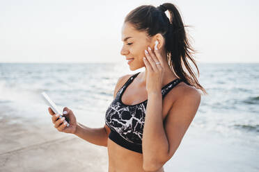 Lächelnde Frau mit drahtlosen In-Ear-Kopfhörern und Smartphone am Strand - OYF00700