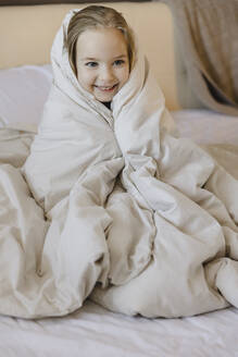 Lächelndes süßes blondes Mädchen, eingewickelt in eine Decke, sitzt auf dem Bett zu Hause - SEAF00619