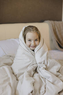 Smiling cute girl wrapped in blanket sitting on bed at home - SEAF00620