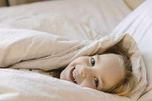 Lächelndes, in eine Decke eingewickeltes Mädchen, das zu Hause auf dem Bett liegt - SEAF00621