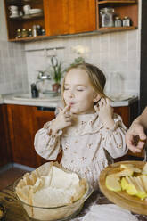 Cute girl with eyes closed tasting food in kitchen at home - SEAF00637