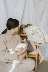Woman sitting with cat looking at daughter climbing wooden steps at home - SEAF00646