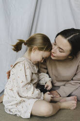 Frau mit geschlossenen Augen küsst ihre Tochter, die mit der Kamera zu Hause sitzt - SEAF00651