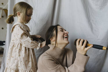 Cheerful woman holding hairbrush singing by daughter tying hair at home - SEAF00654