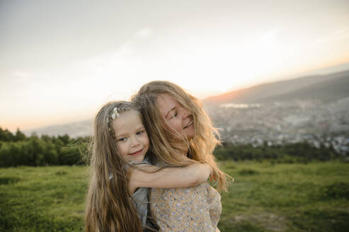 Nettes Mädchen mit blondem Haar umarmt Mutter bei Sonnenuntergang - VBUF00040
