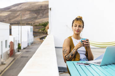 Junge Frau hält Teetasse und sitzt mit Laptop auf dem Dach - MRAF00862