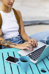 Junge Frau mit Tätowierung tippt auf einem Laptop und sitzt am Tisch - MRAF00863
