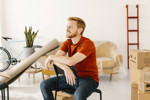 Smiling man sitting on chair in living room at home - XLGF02794