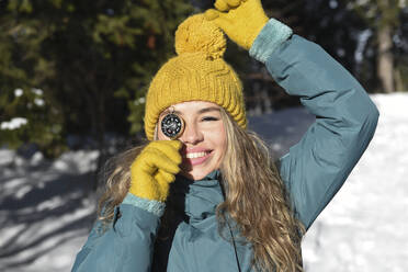 Lächelnde Frau mit gelber Strickmütze, die das Auge mit einem Navigationskompass bedeckt, an einem sonnigen Tag - VBUF00050