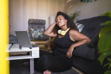 Smiling young woman with hand in hair sitting by sofa in living room - SNF01591