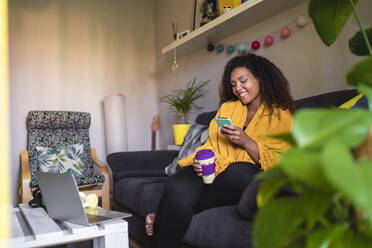 Happy woman holding disposable coffee cup using smart phone sitting on sofa in living room - SNF01600