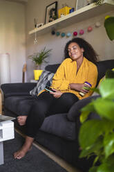 Smiling woman holding disposable coffee cup and smart phone sitting with eyes closed on sofa at home - SNF01602