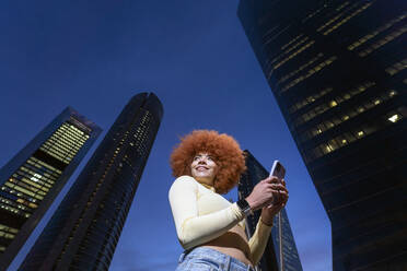 Smiling woman with Afro hairstyle using smart phone in front of skyscrapers - OCMF02337