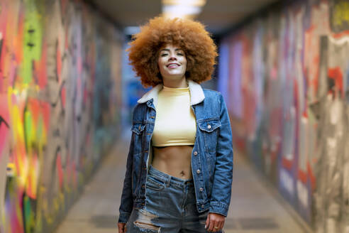 Smiling woman with Afro hairstyle standing by graffiti wall - OCMF02338