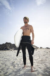 Mann ohne Hemd mit Surfbrett am Strand stehend - FBAF01830