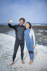 Glückliches Paar im Neoprenanzug, das ein Selfie mit seinem Smartphone am Strand macht - FBAF01836