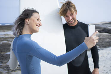 Glückliches Paar im Neoprenanzug macht Selfie mit Surfbrett am Strand - FBAF01837