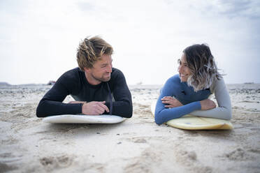 Glückliches Paar im Neoprenanzug auf einem Surfbrett am Strand liegend - FBAF01840