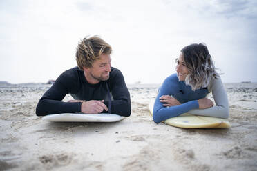 Happy couple in wetsuit lying down on surfboard at beach - FBAF01840